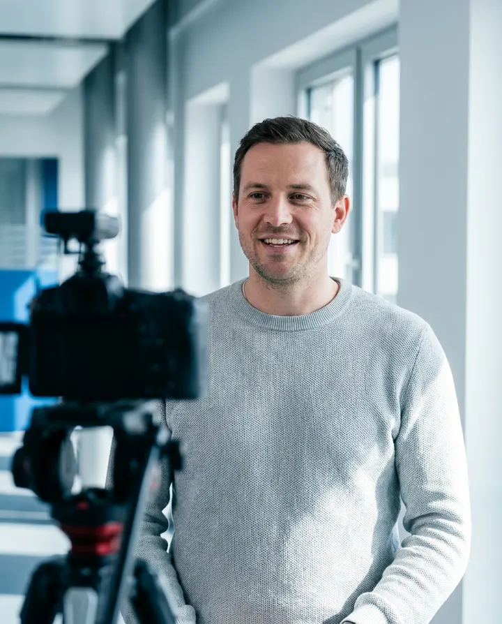 A presenter in a grey sweater talking to camera in a bright office, a frame from a brand account video